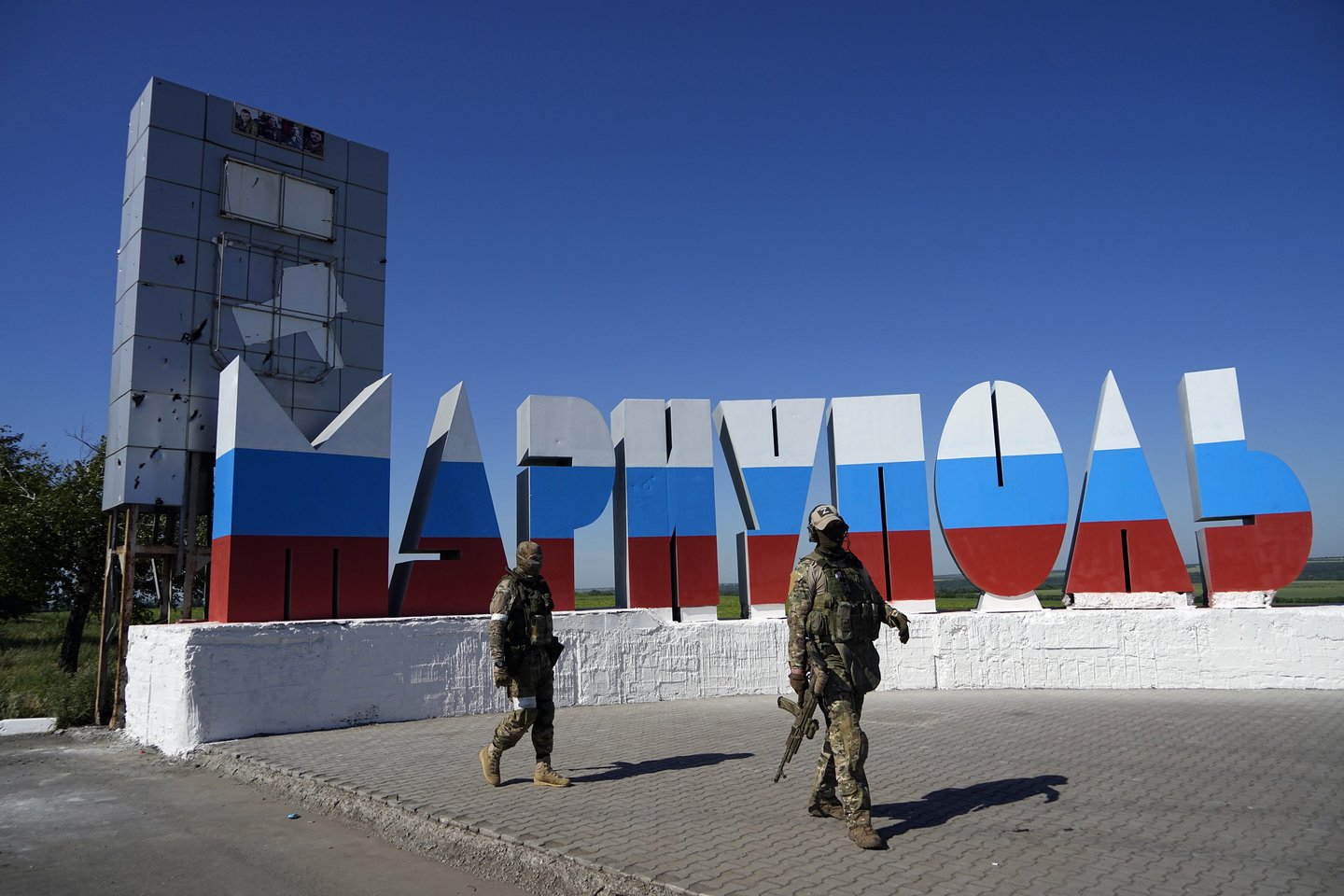 Rusijos kariai eina pro Rusijos vėliavos spalvomis perdažytą miesto pavadinimą prie įvažiavimo į Mariupolį, apsišaukėliškos Donecko liaudies respublikos vyriausybės kontroliuojamoje teritorijoje, Rytų Ukrainoje, sekmadienį, 2022 m. birželio 12 d.<br>AP/Scanpix nuotr.